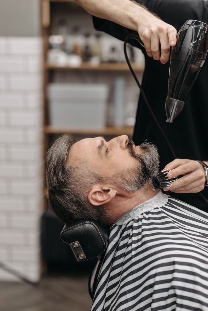 Man receiving beard styling at barber shop with blow dryer and brush.